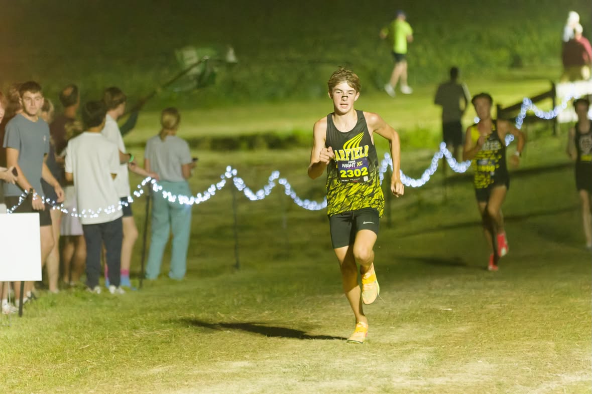 A Garfield cross country runner nears the finish line at a night meet lit by string lights, with other runners close behind